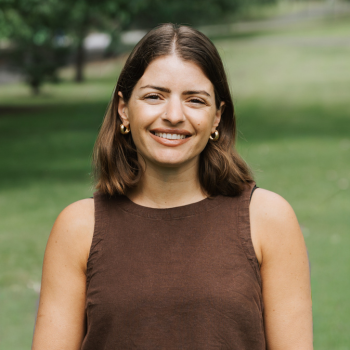 Outdoor headshot of a smiling woman with shoulder-length brown hair, wearing a sleeveless brown top.